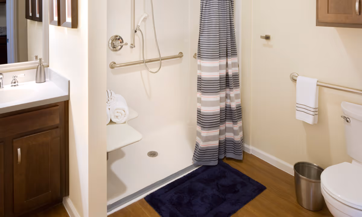 A clean and accessible bathroom featuring a walk-in shower with a striped shower curtain, a built-in shower seat with rolled white towels, grab bars, a wooden vanity with a white countertop and soap dispenser, a toilet with a towel hanging on a grab bar, a small trash can, and a dark blue bath mat on a wooden floor.