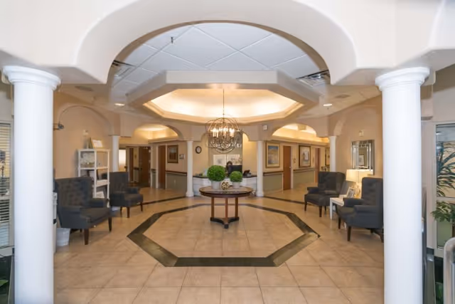 Well-lit reception lobby with a central round table under a chandelier, columns, seating alcoves and tiled floor.