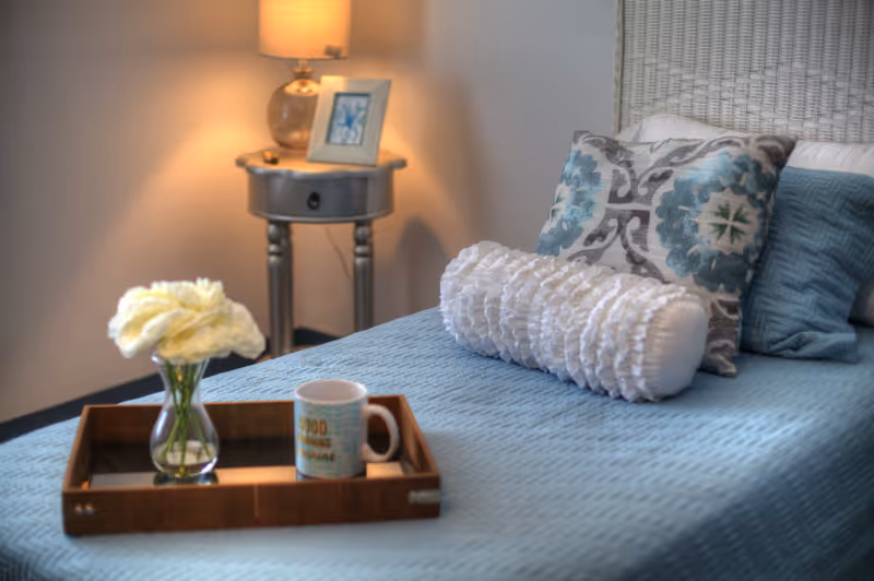 A neatly made bed with a light blue quilted bedspread, decorative pillows including a white ruffled bolster pillow and patterned cushions. On the bed is a wooden tray holding a small vase with white flowers and a coffee mug. In the background, there is a round bedside table with a lamp and a framed picture.