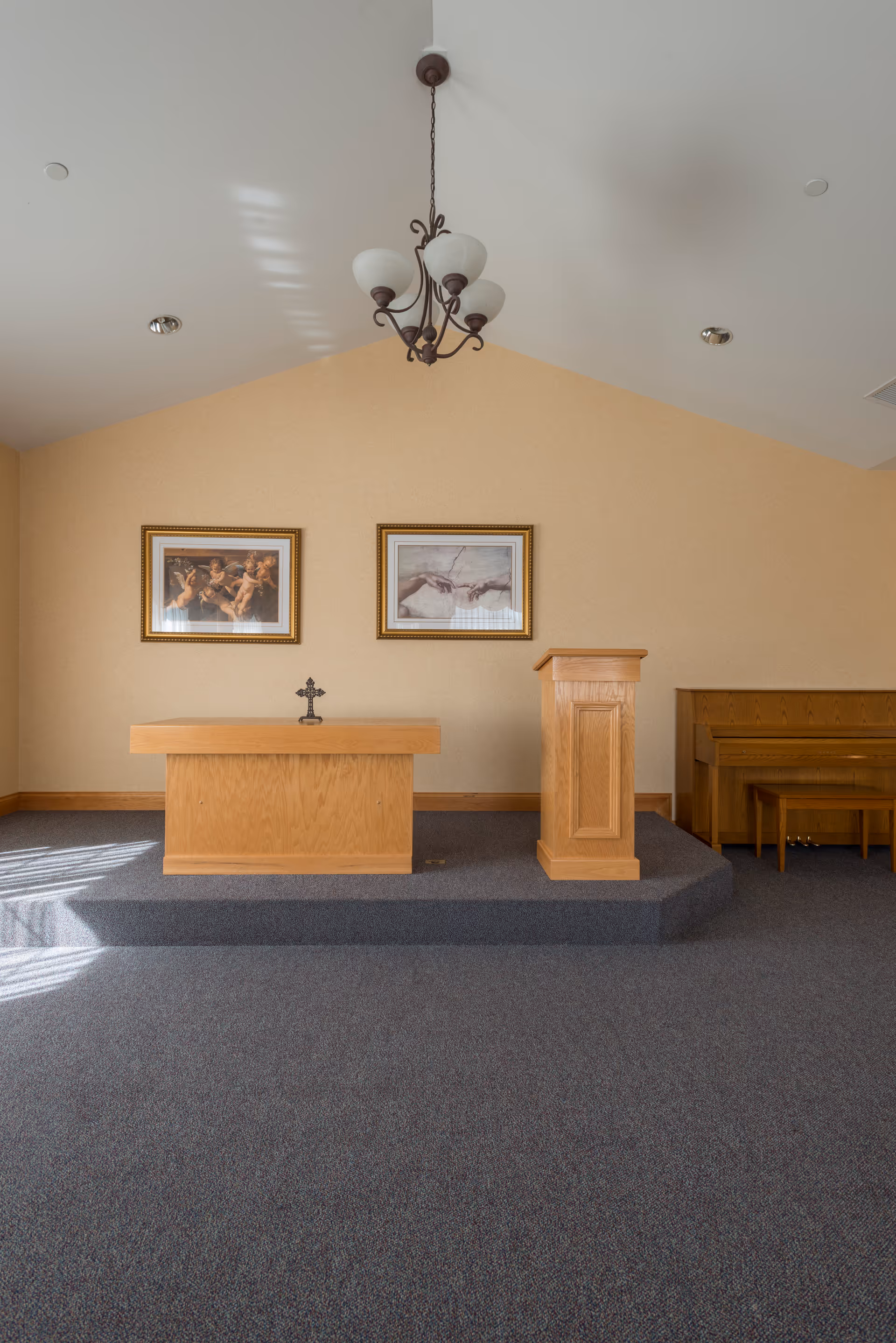 A small chapel or prayer room with a raised platform featuring a wooden altar with a cross, a wooden lectern, and a piano with a bench to the right. Two framed religious paintings hang on the beige wall behind the altar. A chandelier with five lights hangs from the ceiling.