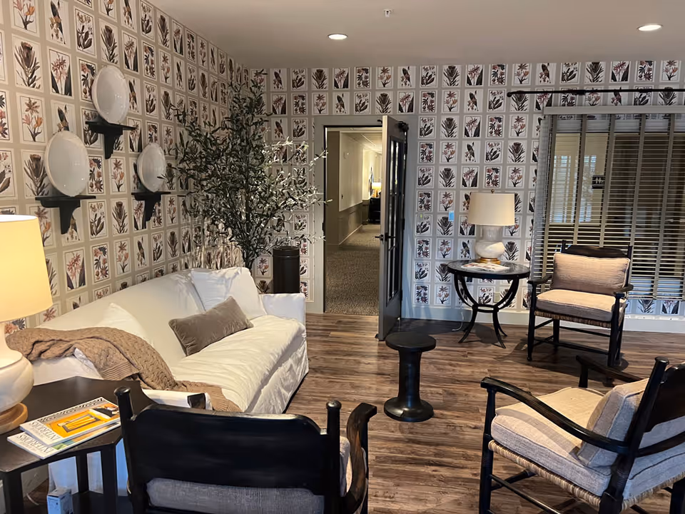 A cozy living room with a white couch, three cushioned wooden chairs, a small round table with a lamp, and a plant in the corner. The walls are decorated with framed botanical prints arranged in a grid pattern. There is a door open leading to a hallway and a window with blinds on the right side.