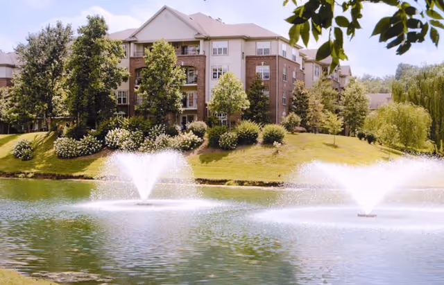A multi-story brick residential building surrounded by trees and landscaping, viewed from across a pond with two water fountains spraying water.