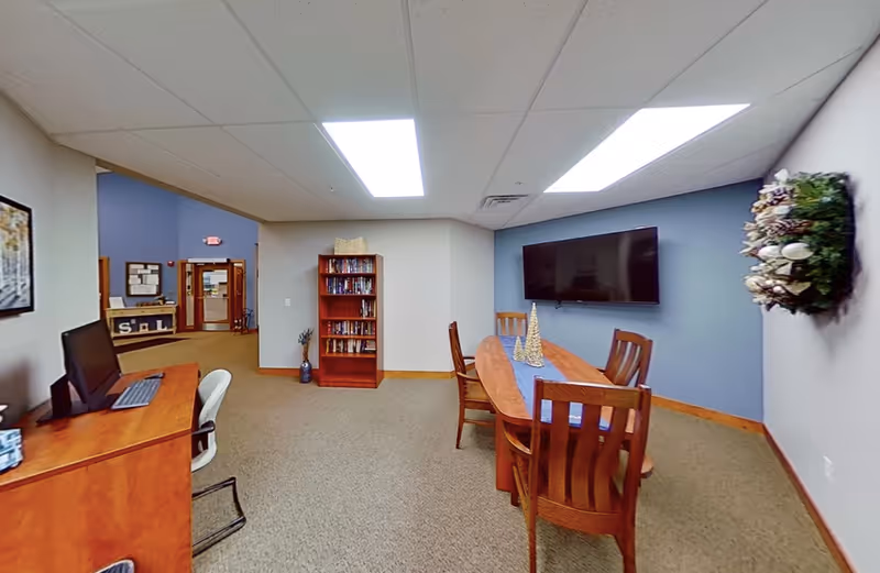 A senior living facility common area with a wooden table and four chairs, a flat-screen TV mounted on a blue accent wall, a decorative wreath on the adjacent wall, a wooden bookshelf filled with books, and a desk with a computer and chair. The room has beige carpet and white ceiling tiles with fluorescent lighting.