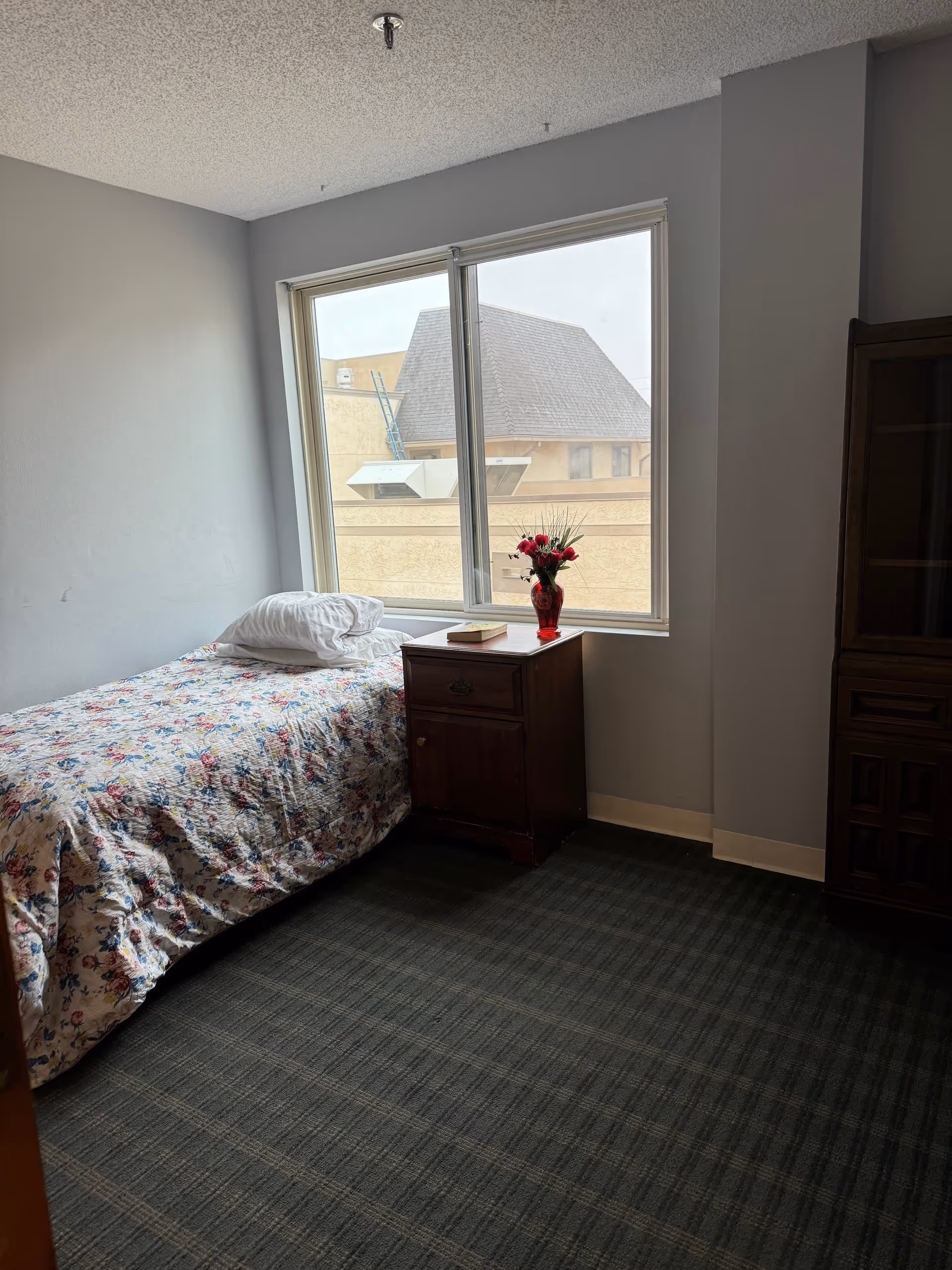 Sunlit small bedroom with a single bed covered in a floral quilt, a wooden nightstand holding a vase of red flowers beneath a large window.