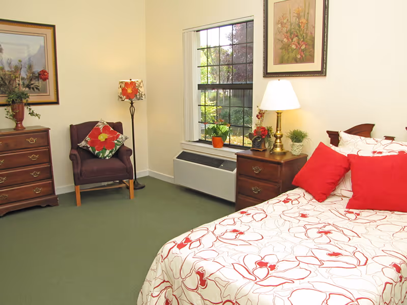 A cozy bedroom with a bed covered in white and red floral bedding, two red pillows, a wooden nightstand with a lamp and small plants, a window showing greenery outside, a purple armchair with a floral cushion, a floor lamp, and a wooden dresser with a vase of flowers and framed artwork on the walls.
