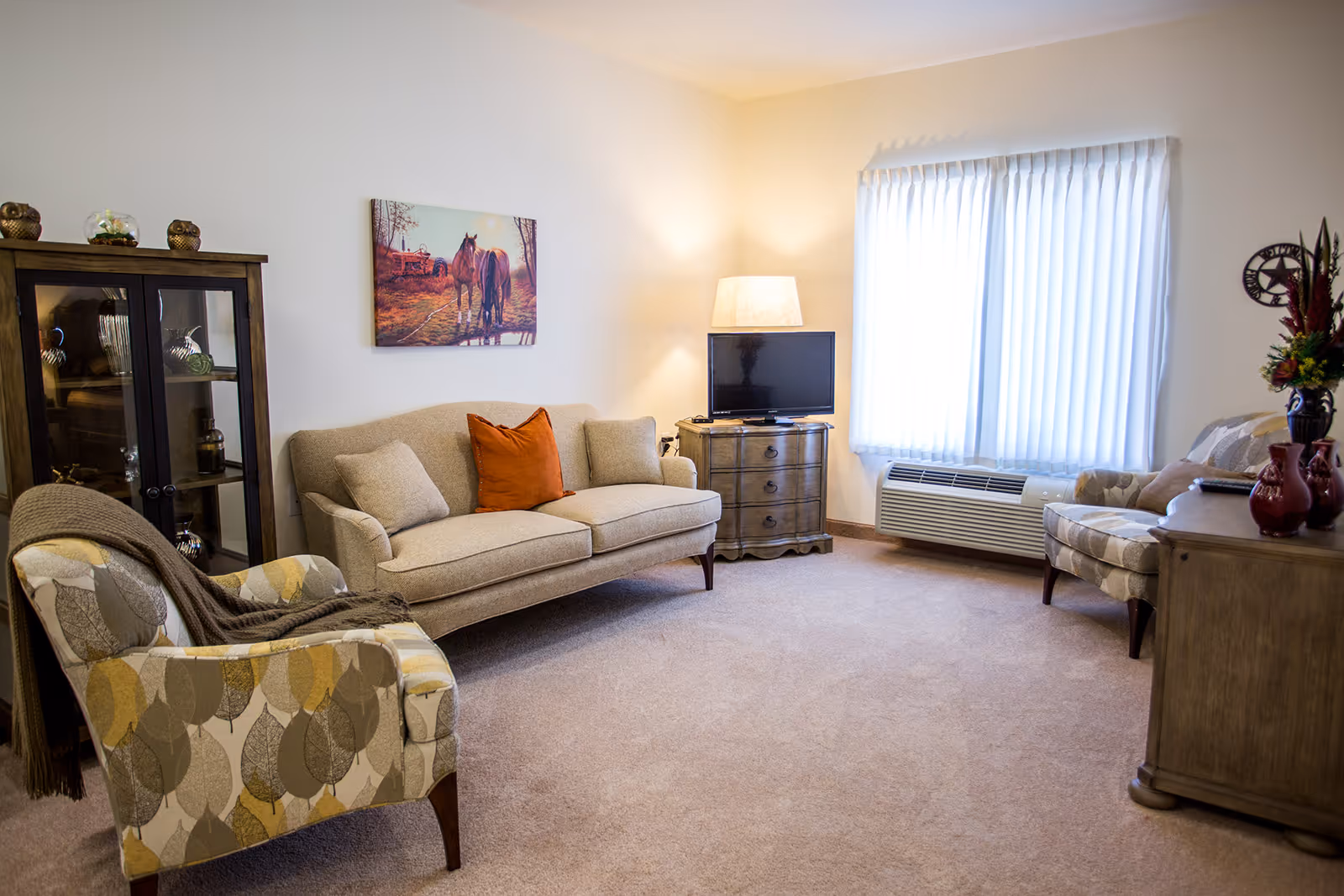 A cozy living room with a beige sofa adorned with two beige pillows and one orange pillow, two patterned armchairs, a wooden cabinet with glass doors displaying decorative items, a small wooden dresser with a flat-screen TV and a lamp on top, a window with white vertical blinds, and a carpeted floor.