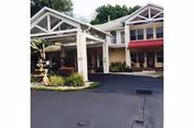Exterior view of Bayou Gardens Assisted Living facility showing the entrance with a covered driveway, landscaped greenery, and a two-story building with windows and a red awning.
