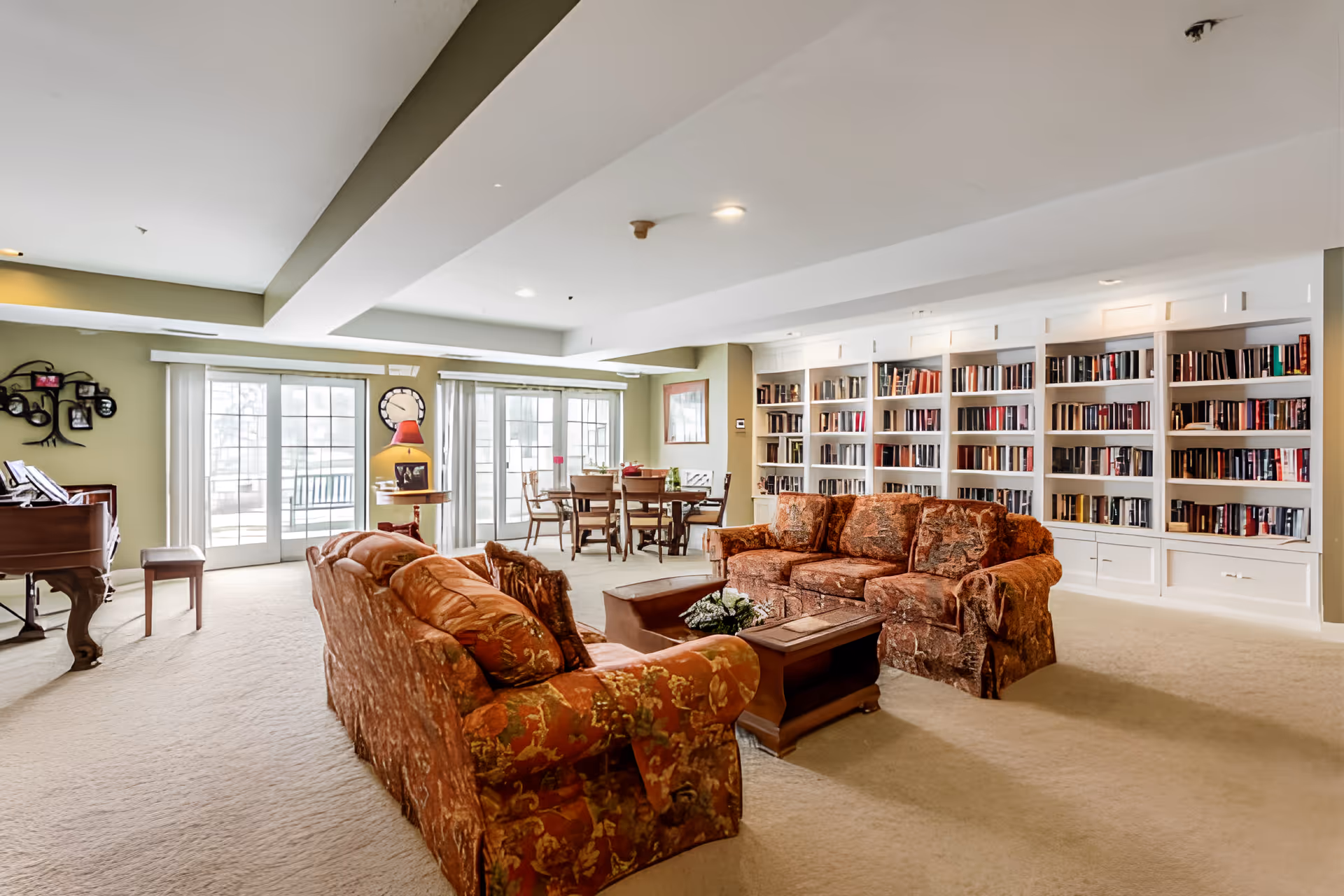 A spacious living room area in Elison Independent Living of Orchard Glen featuring two patterned sofas facing each other with a wooden coffee table in between. Behind the sofas is a large white built-in bookshelf filled with books. To the left, there is a grand piano and a small bench. In the background, there is a dining table with chairs near large sliding glass doors that let in natural light. The room has a beige carpet and light-colored walls with some decorative items like a wall clock and framed pictures.