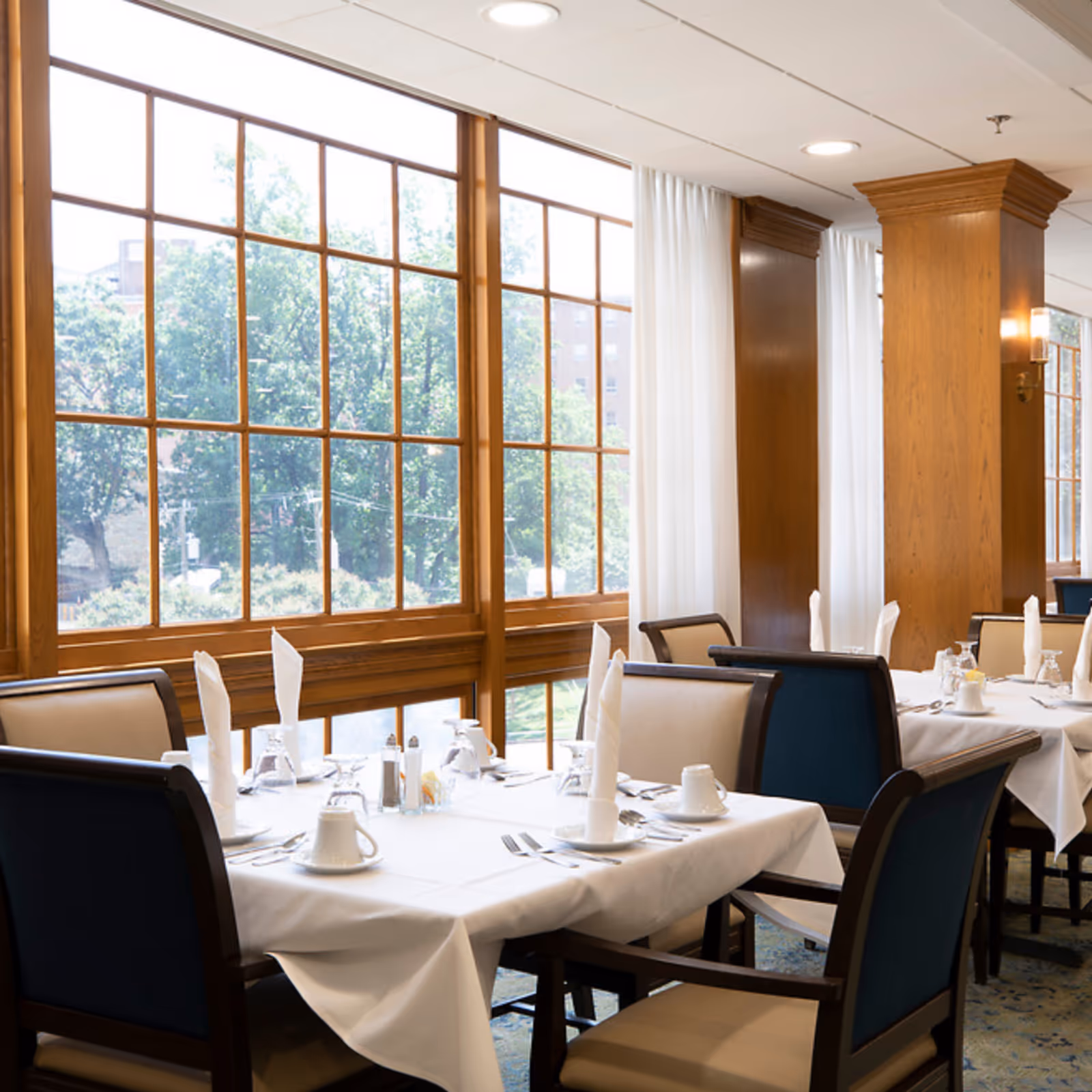 A bright dining room with large wooden-framed windows letting in natural light. Tables are set with white tablecloths, folded white napkins, cups, glasses, and silverware. The room features wooden columns and light-colored curtains.