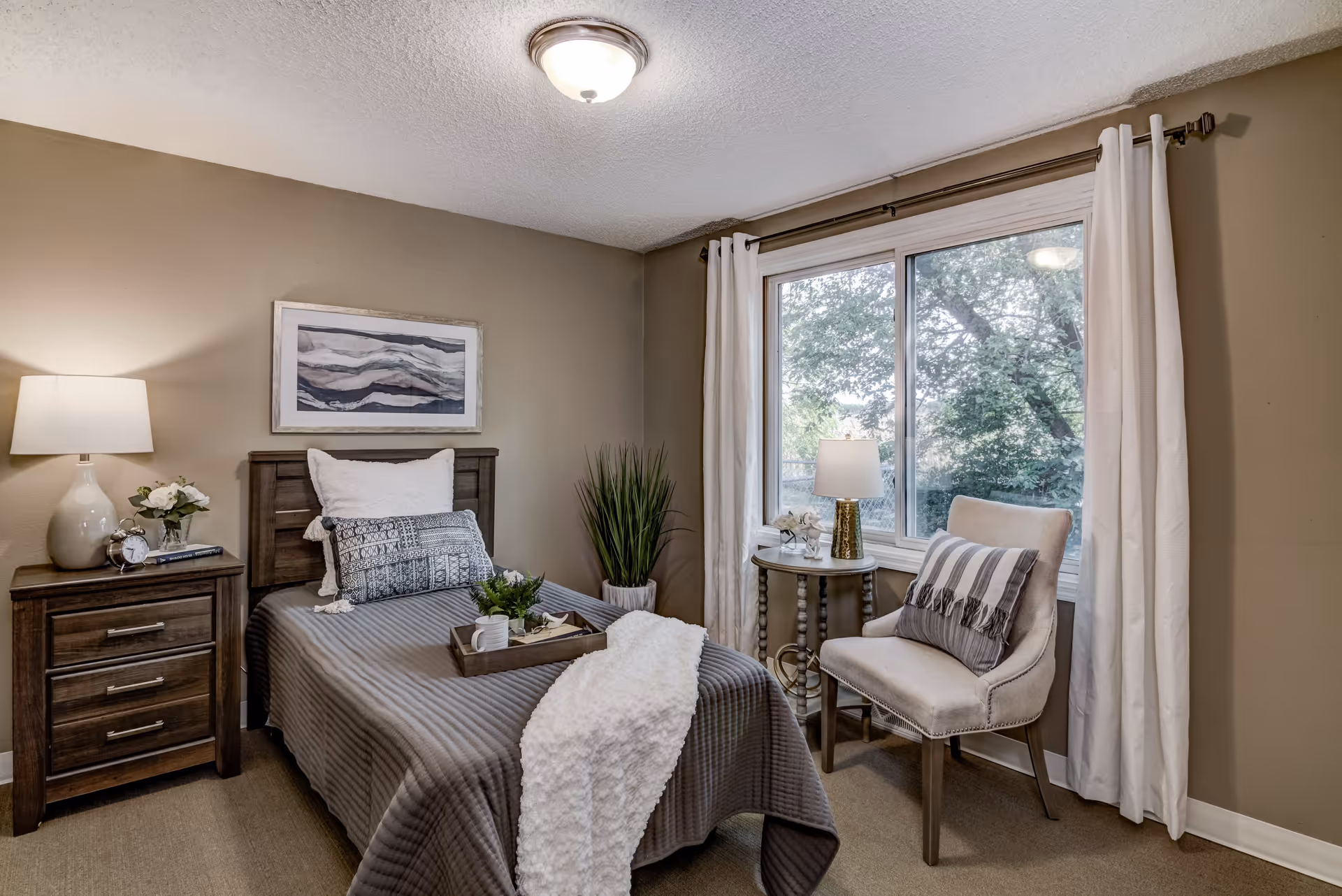A cozy bedroom with a single bed covered in a gray quilt and white throw blanket. There is a wooden nightstand with a lamp, clock, and flowers on the left side of the bed. A framed abstract artwork hangs above the bed. To the right, there is a window with white curtains, a small round table with a lamp and flowers, and a beige upholstered chair with a striped pillow.