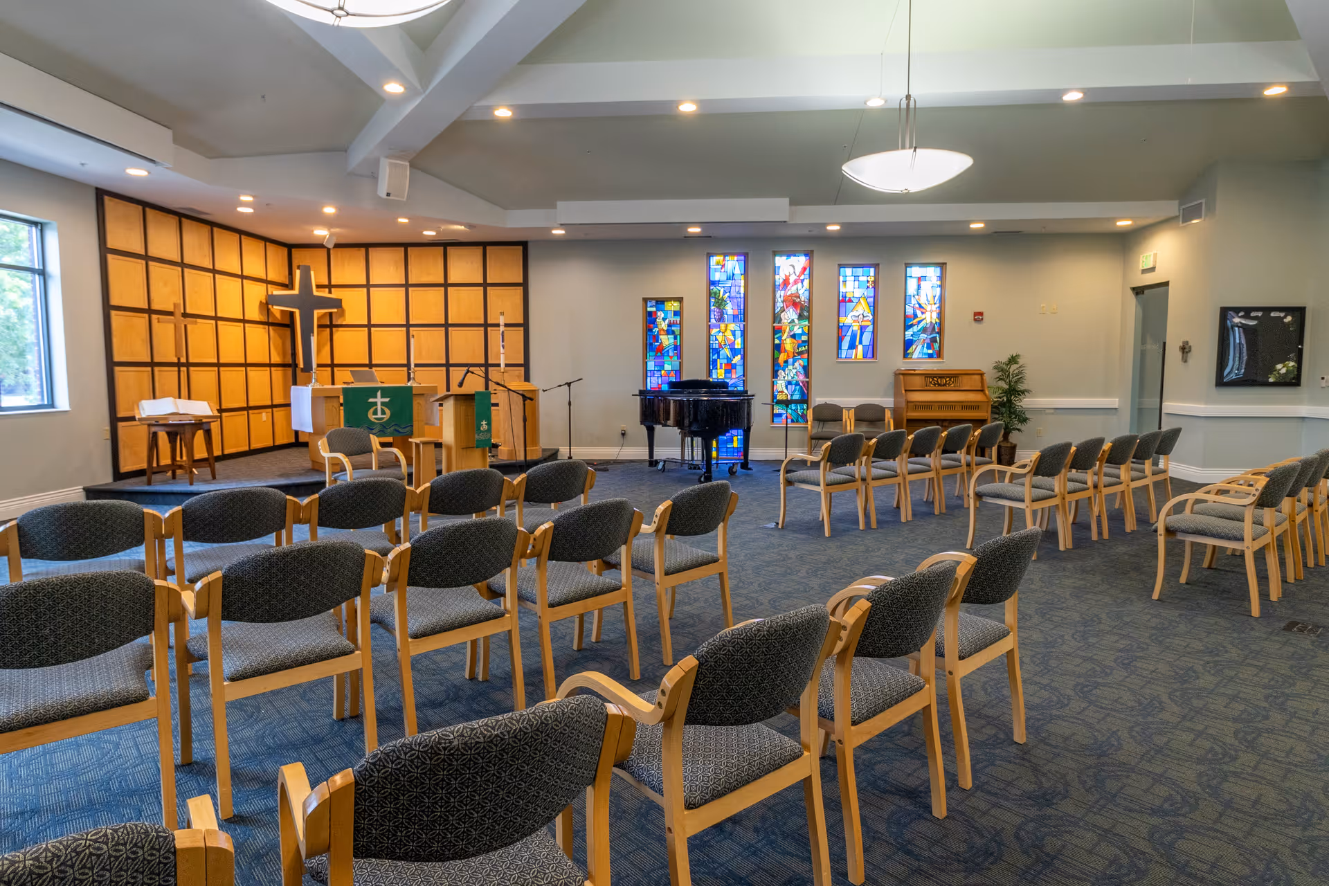 Chapel-style meeting room with rows of chairs facing an altar, a large cross, a piano, and stained glass windows.
