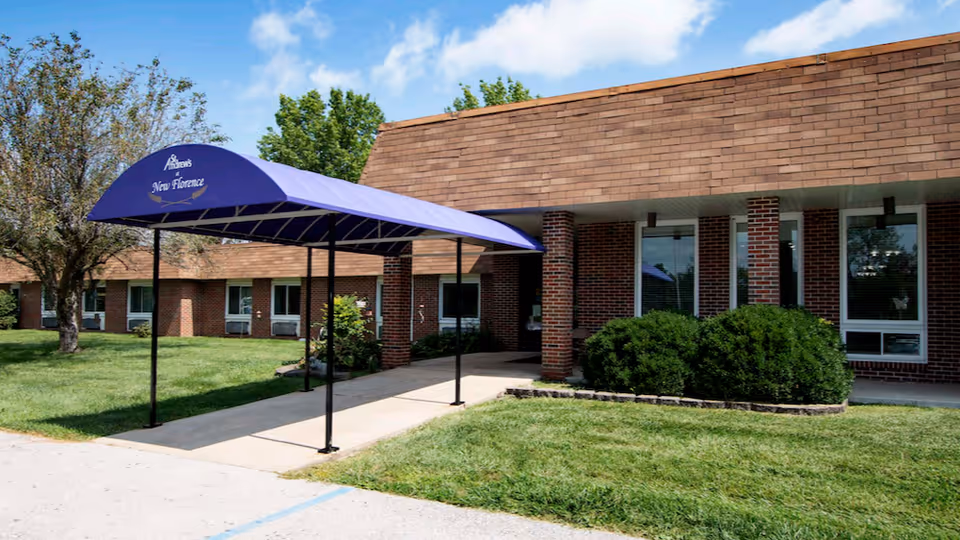 Exterior view of St. Andrew's at New Florence facility showing a brick building with a covered entrance canopy in purple. The canopy has the facility name printed on it. There is a well-maintained lawn with bushes and trees under a partly cloudy blue sky.