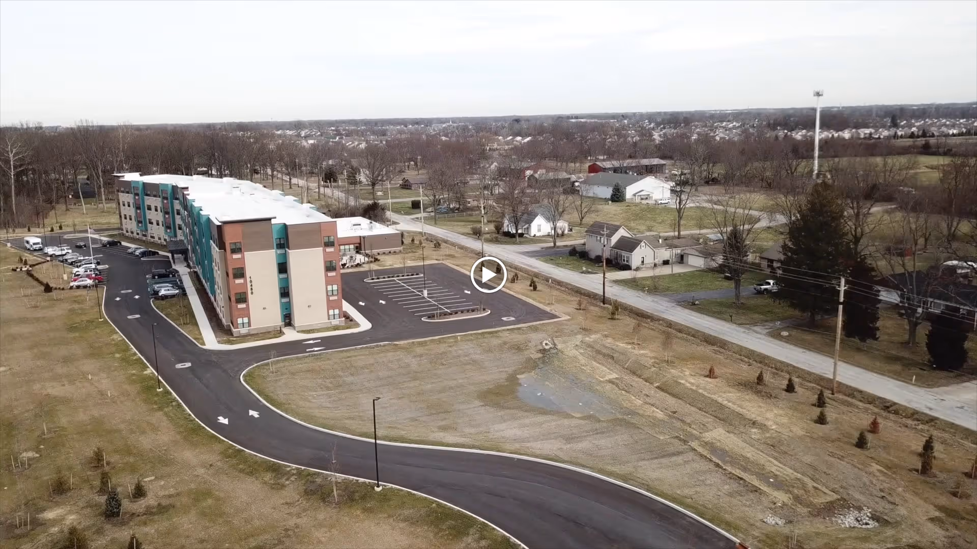 Aerial view of a multi-story senior living facility building with a parking lot and surrounding grassy area. The building is situated near a road with residential houses and trees in the background under a clear sky.