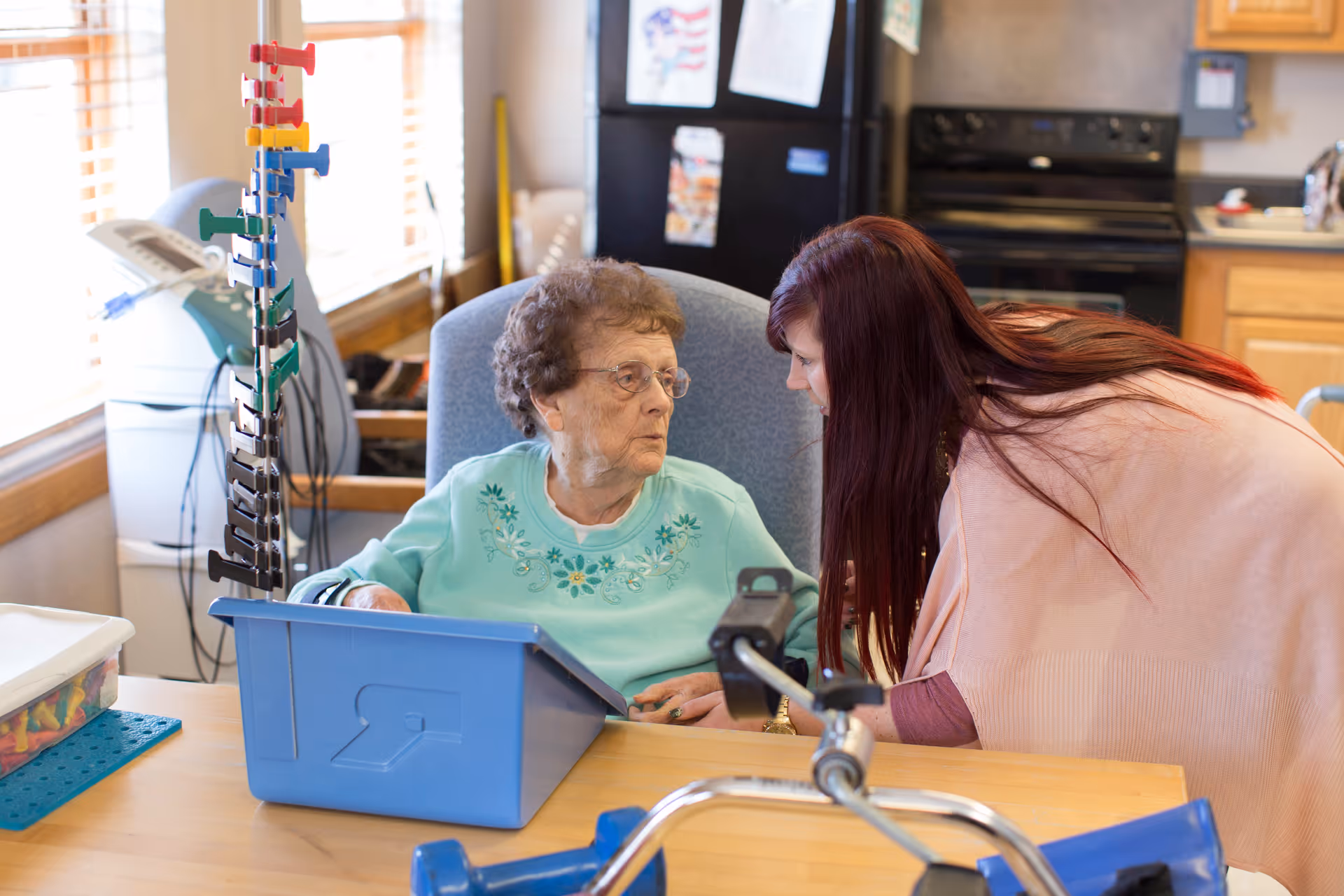 An elderly woman sitting in a chair at a table with a blue plastic bin in front of her, interacting with a younger woman who is leaning towards her. The setting appears to be a kitchen or common area with a refrigerator, stove, and cabinets in the background.