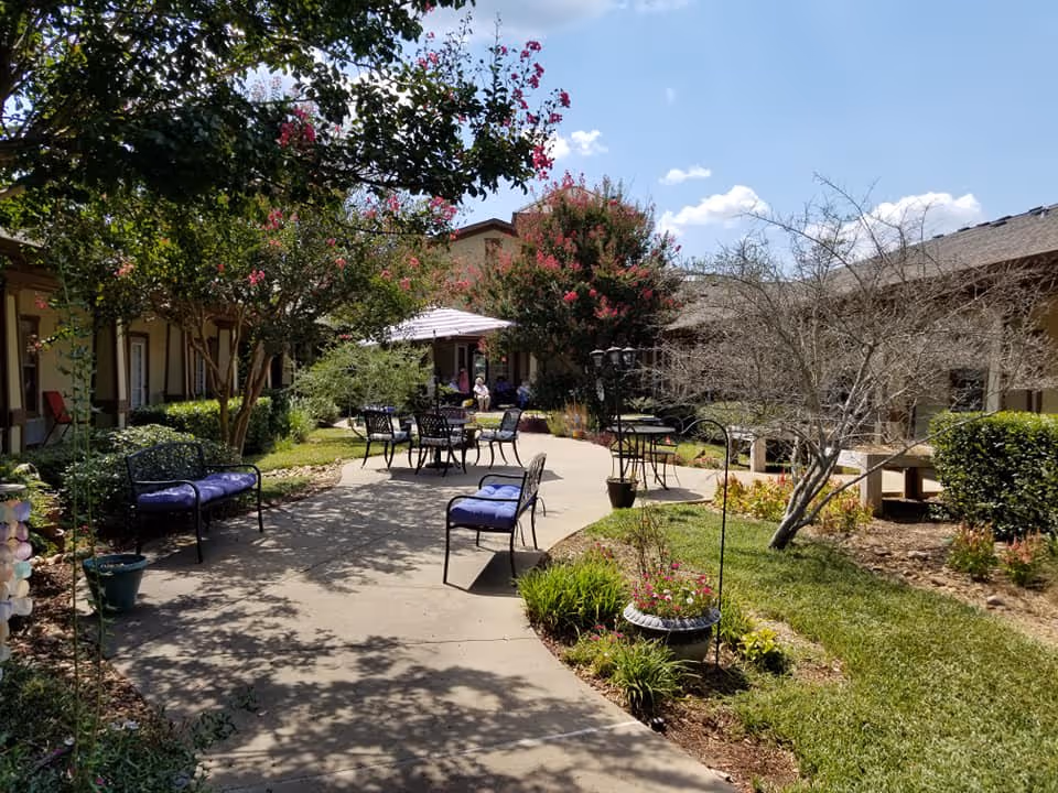 Outdoor courtyard area with a paved walkway surrounded by green grass, flowering trees, and shrubs. Several black metal chairs and tables with blue cushions are arranged along the path. The sky is clear with a few clouds, and the buildings of the assisted living facility are visible in the background.