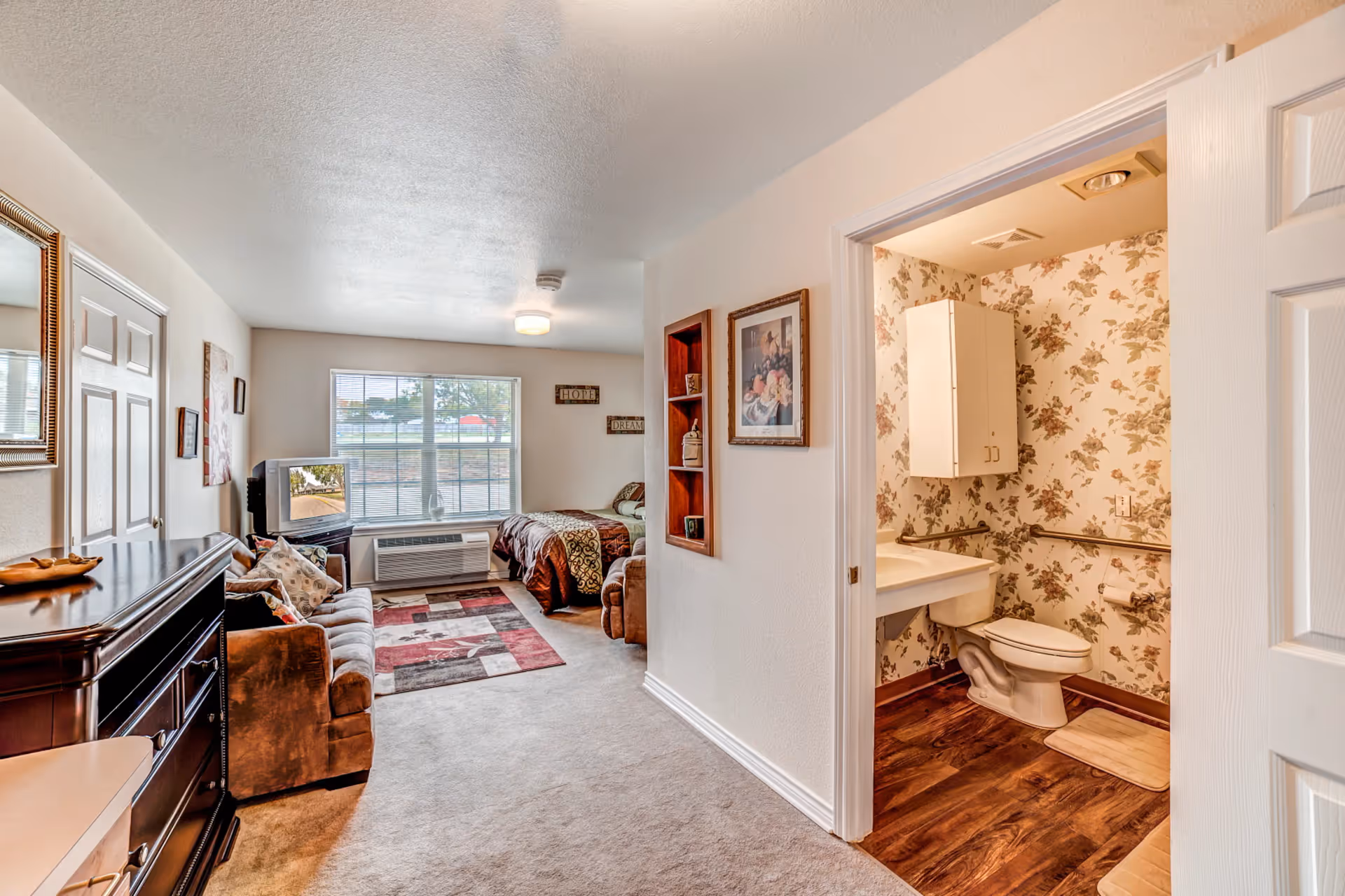 Interior view of a senior living facility room featuring a living area with a brown couch, a wooden dresser, a TV on a stand, a bed near a large window with blinds, and a bathroom with floral wallpaper, a toilet, and a sink visible through an open door.
