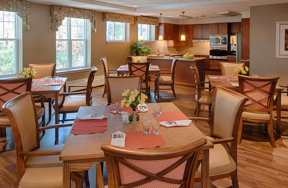 A cozy dining room in a senior living facility with several wooden tables and chairs arranged neatly. Each table is set with placemats, napkins, glasses, and small flower vases. Large windows with floral valances let in natural light, and a kitchen area with wooden cabinets and modern appliances is visible in the background.