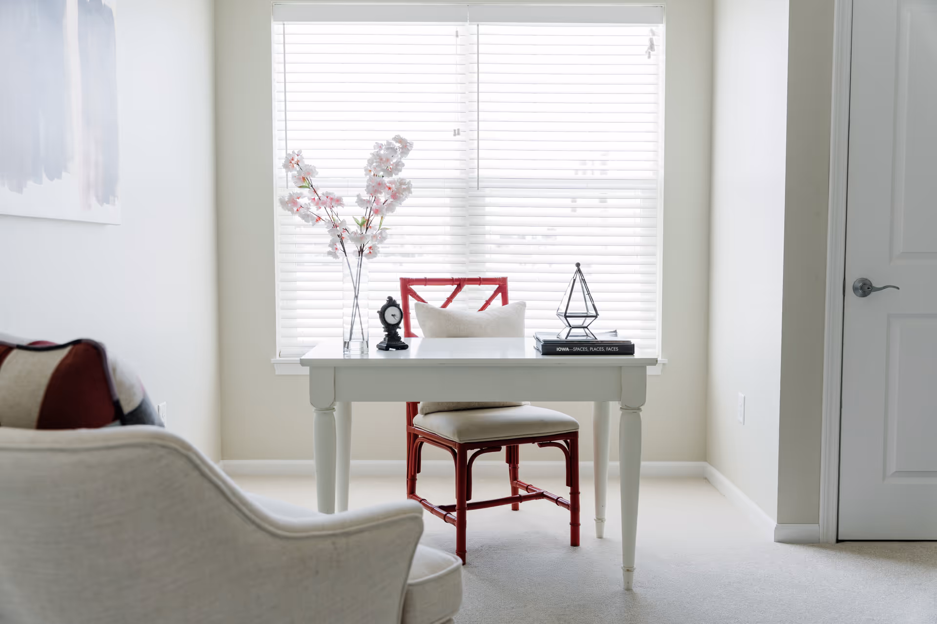 A bright room with a white desk and a red chair placed in front of a window with white blinds. On the desk, there is a vase with pink flowers, a small black clock, a geometric decorative object, and a stack of books. A beige cushioned armchair is partially visible in the foreground, and a closed white door is on the right side of the room.