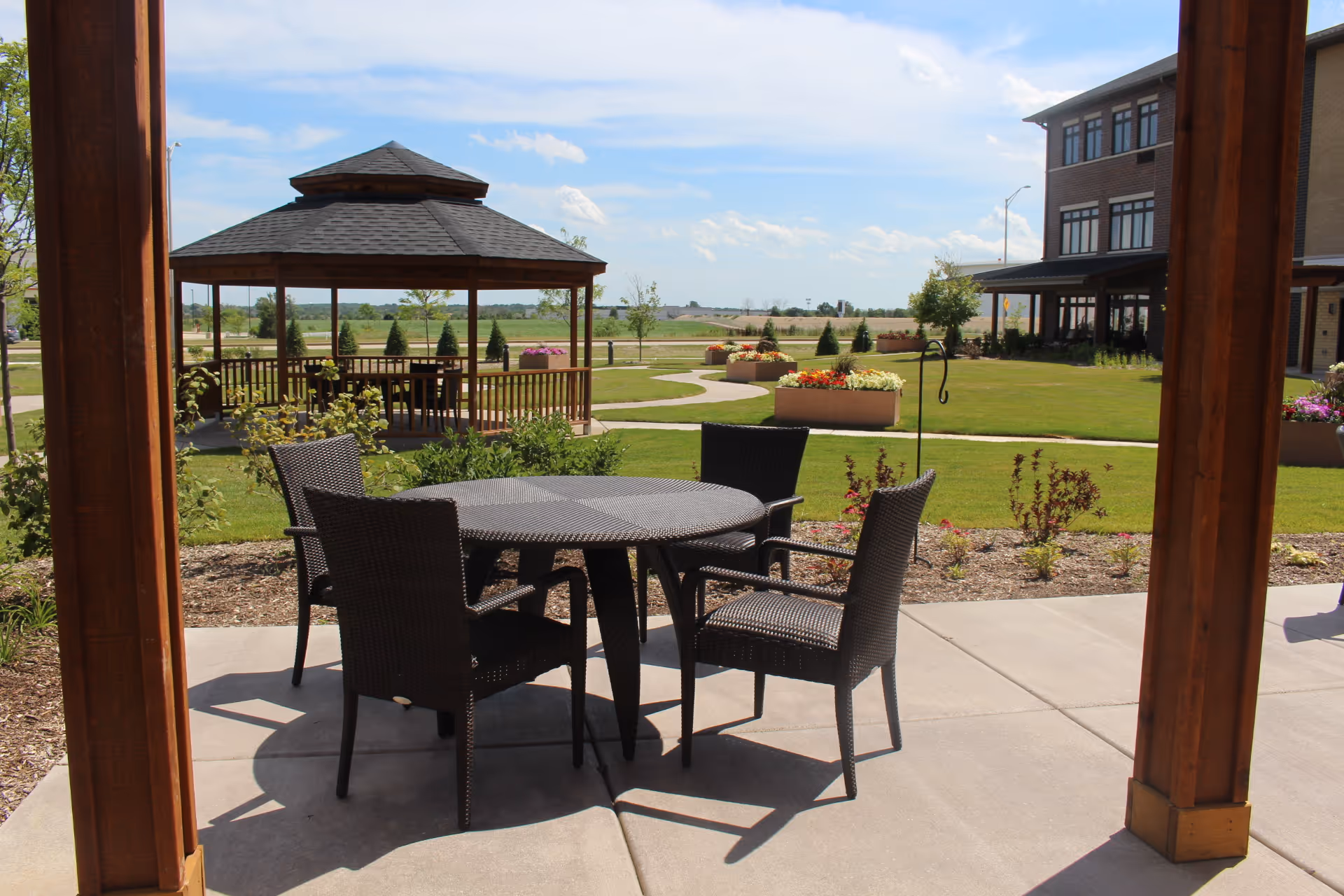 Outdoor patio area with a round table and four wicker chairs under a wooden pergola. In the background, there is a gazebo, green lawn, flower beds, and a multi-story building under a partly cloudy sky.