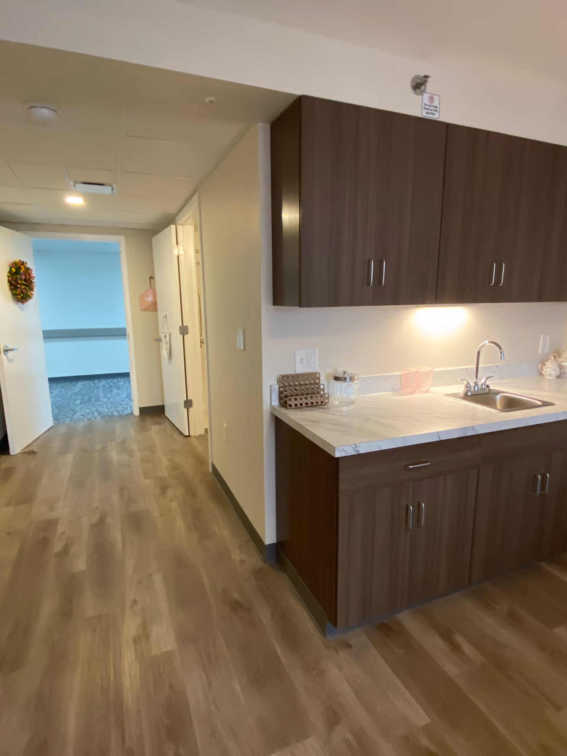 Interior view of a senior living facility showing a hallway with wood flooring, a kitchen counter with a sink and dark wood cabinets on the right, and an open door decorated with a wreath leading to another room with carpeted flooring.