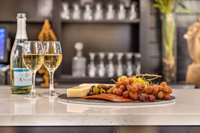 A close-up view of a marble countertop with a charcuterie board featuring grapes, cheese, olives, and sliced meat. Two glasses of white wine and a bottle of sparkling water are placed next to the board. In the background, shelves with glassware and decorative items are visible.