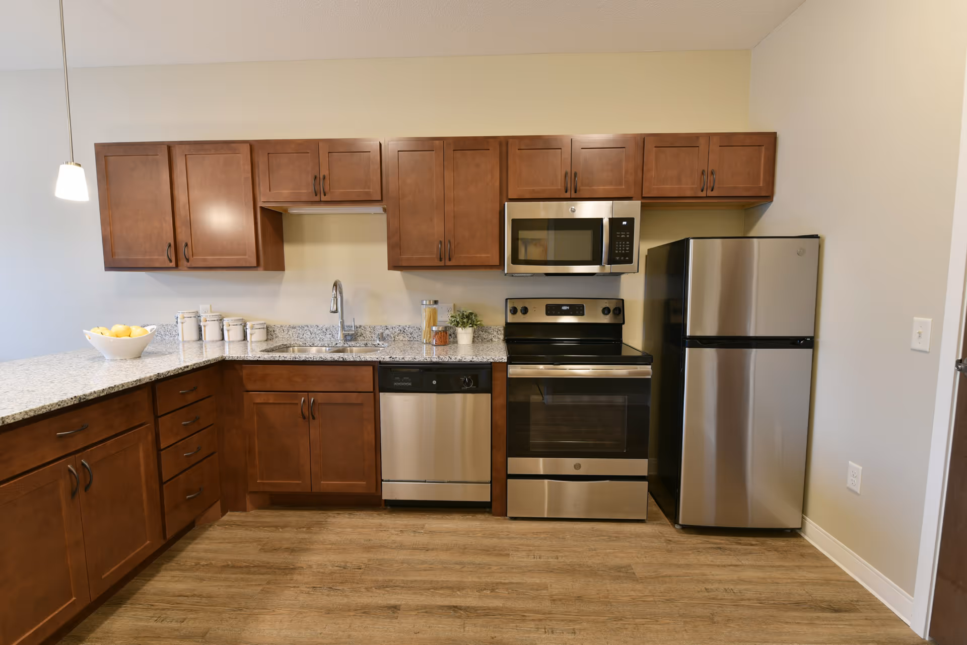 Modern kitchen with wooden cabinets, granite countertops, stainless steel appliances including a refrigerator, oven, microwave, and dishwasher. There is a bowl of lemons and some jars on the counter, and a small plant next to the stove.