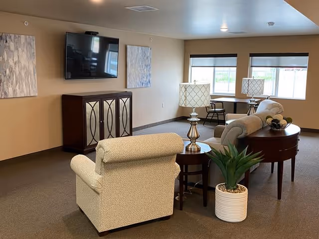 A cozy living room area in an assisted living facility featuring a beige armchair, a sofa, two side tables with decorative lamps, a potted plant, a TV mounted on the wall above a wooden cabinet, two abstract paintings on the wall, and a window with a desk and chair in the background.