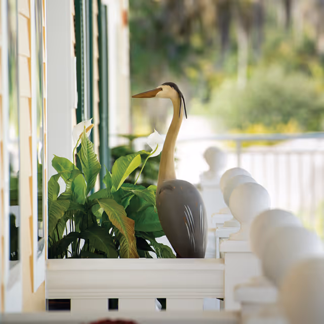 A decorative bird statue resembling a heron stands among green leafy plants on a white porch railing with blurred outdoor greenery in the background.