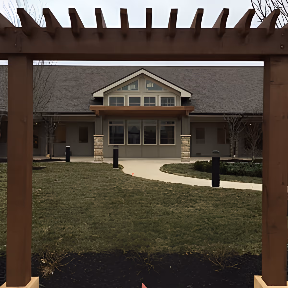 Front exterior of a senior living facility framed by a wooden pergola, with a grassy courtyard and curved walkway.
