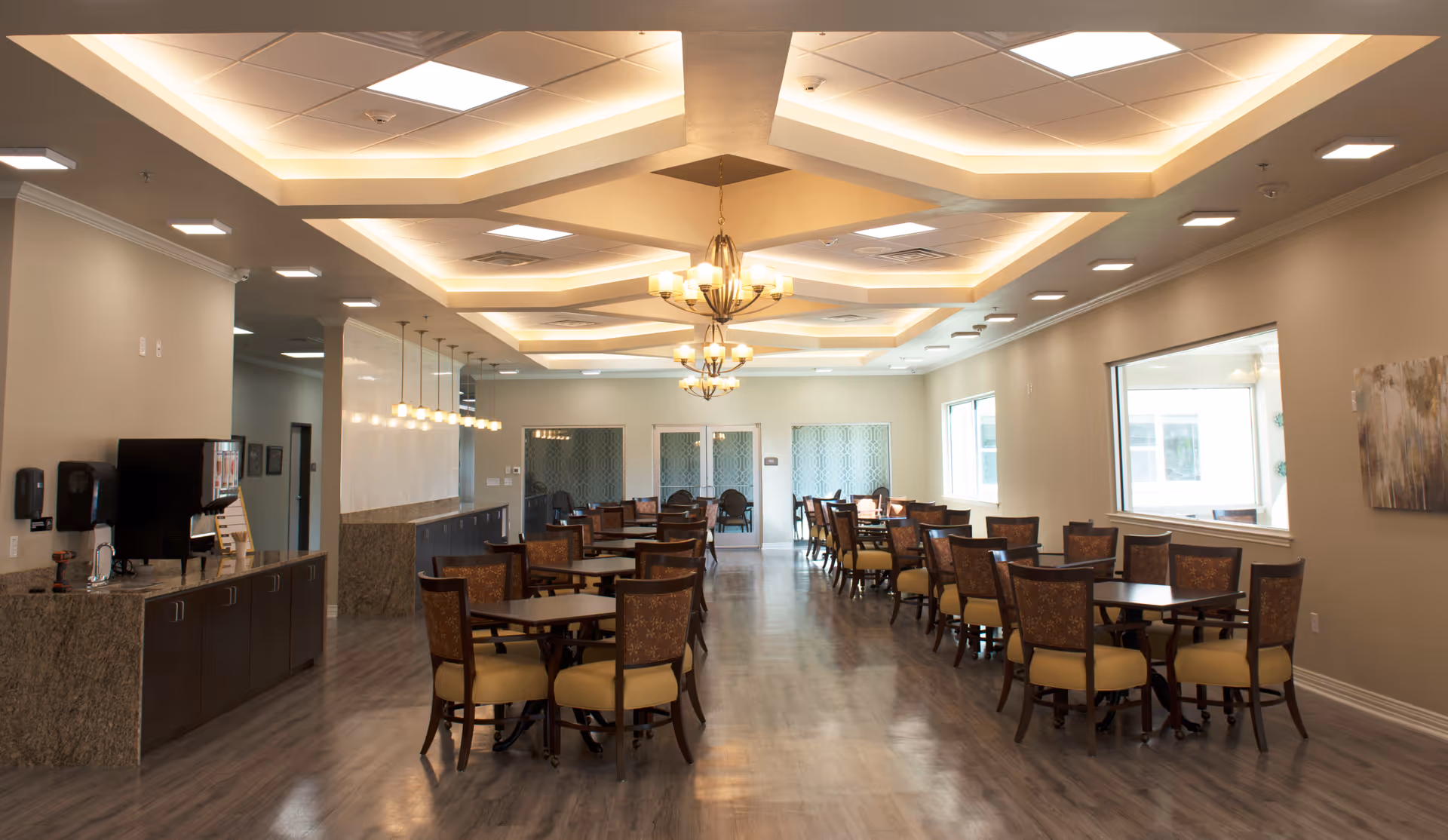 A spacious dining room with multiple wooden tables and cushioned chairs arranged neatly. The room features a modern ceiling design with recessed lighting and two chandeliers. There is a counter area with a beverage dispenser and sink on the left side, and large windows on the right wall allowing natural light to enter.