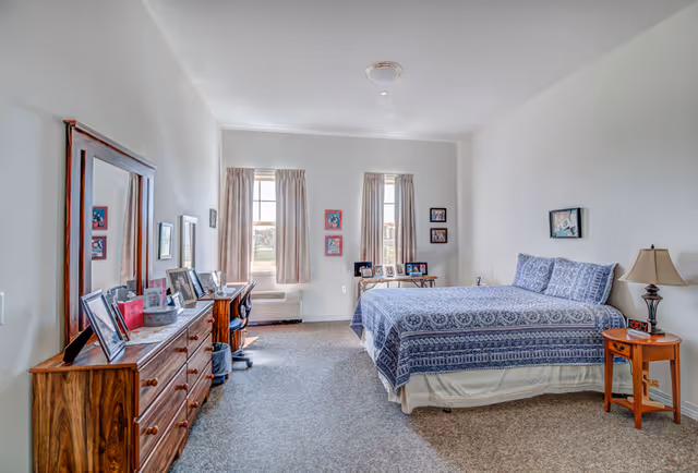 Spacious, well-lit bedroom with a blue-patterned bed, wooden dresser with mirror, desk, and two windows.