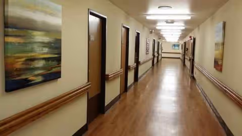 Long well-lit interior hallway in a senior living facility with doors and wall artwork.