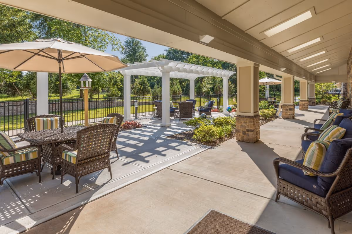 Outdoor patio area at Sunrise of Bridgewater featuring wicker chairs with cushions, tables with umbrellas, a white pergola, and landscaped greenery under a covered walkway with skylights.