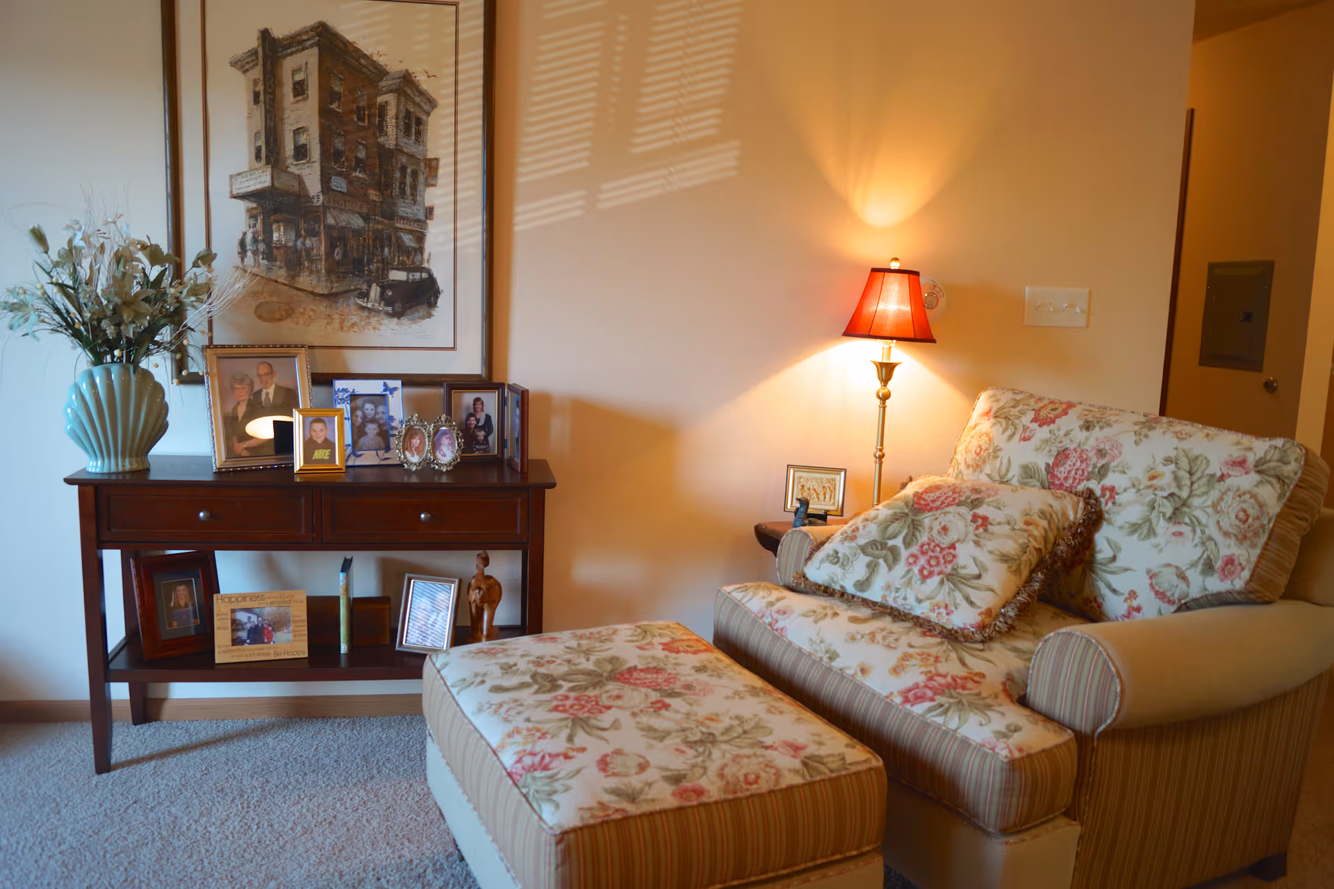 Cozy living room corner with a floral patterned armchair and matching ottoman, a wooden side table with framed family photos and a vase with flowers, a floor lamp with a red lampshade casting warm light on the beige wall, and a framed artwork hanging above the table.
