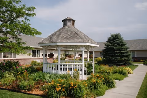A white wooden gazebo with a shingled roof sits in the center of a landscaped garden with various green plants and orange flowers. A paved walkway leads past the gazebo, and a building with beige siding and a gray roof is visible in the background under a partly cloudy sky.