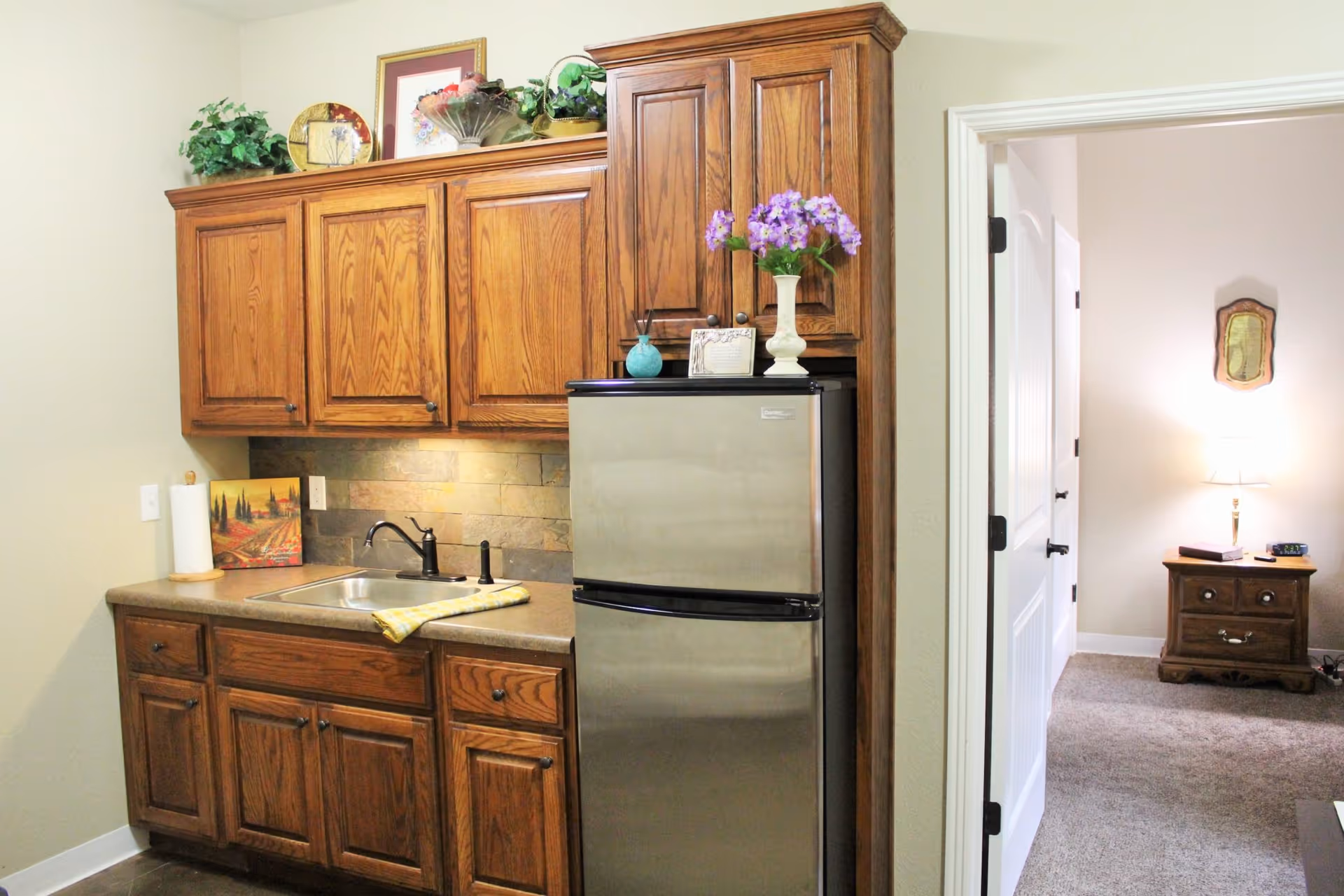 A small kitchen area with wooden cabinets, a stainless steel refrigerator, a sink with a black faucet, and a countertop with a paper towel holder and a decorative painting. On top of the cabinets are plants and framed artwork. To the right, an open door leads to a carpeted room with a wooden nightstand, a lamp, and a wall decoration.