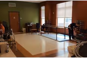 Rehabilitation/exercise room with parallel bars, chairs, walkers and a few people near large windows.