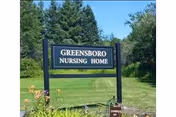 A black sign with white letters reading 'GREENSBORO NURSING HOME' stands on a grassy area with trees and bushes in the background under a clear blue sky.