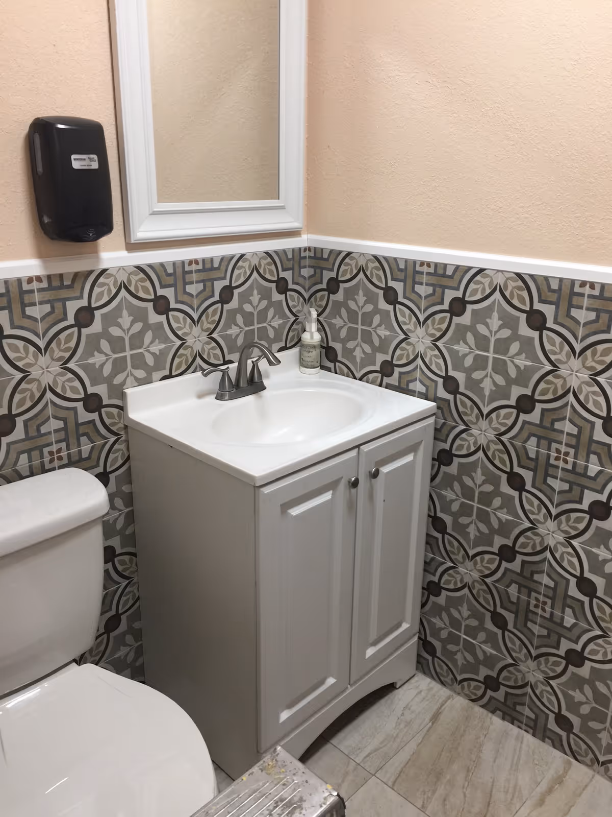 A small bathroom corner featuring a white toilet and a white vanity with a sink and silver faucet. Above the sink is a white-framed mirror and a black soap dispenser mounted on the wall. The walls are decorated with patterned tiles in shades of gray, beige, and white, with a beige painted upper section.