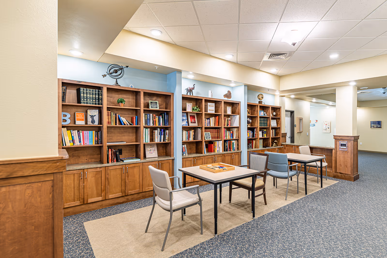 Interior view of a memory care facility room with wooden bookshelves filled with books and decorative items. Two tables with chairs are placed on a beige rug in front of the bookshelves. The room has a carpeted floor, beige walls, and recessed ceiling lights.