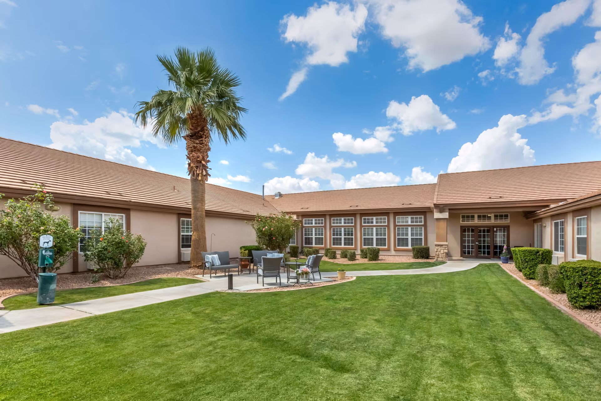 Outdoor courtyard area of a senior living facility with a well-maintained green lawn, a tall palm tree, bushes, and a seating area with chairs and a small table. The building surrounds the courtyard with multiple windows and a tiled roof under a partly cloudy blue sky.