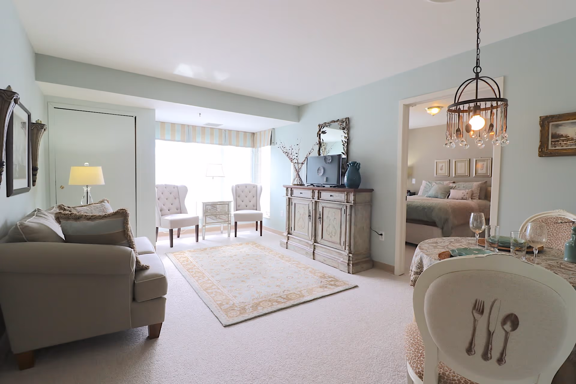 A cozy living room with light blue walls, a beige sofa with pillows, two white armchairs near a large window with striped valance, a decorative rug on the carpeted floor, a vintage-style wooden cabinet with a TV and vase on top, and a dining area with a round table set with plates, glasses, and a chandelier above. A bedroom is visible through an open doorway.