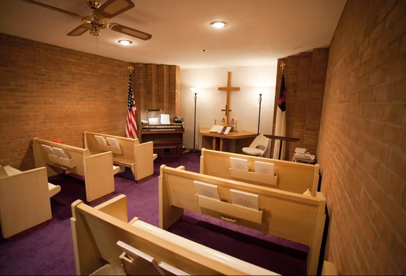 Small chapel room with wooden pews arranged in rows on a purple carpet. At the front, there is a wooden altar with a cross on the wall behind it, two floor lamps, an American flag, a Christian flag, and a small organ. The walls are made of brick.