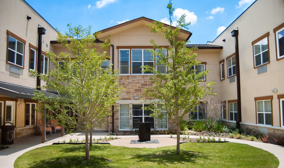 Outdoor courtyard area of a senior living facility with a green lawn, two small trees, a central black sculpture, and a two-story beige building with multiple windows surrounding the courtyard under a blue sky with some clouds.