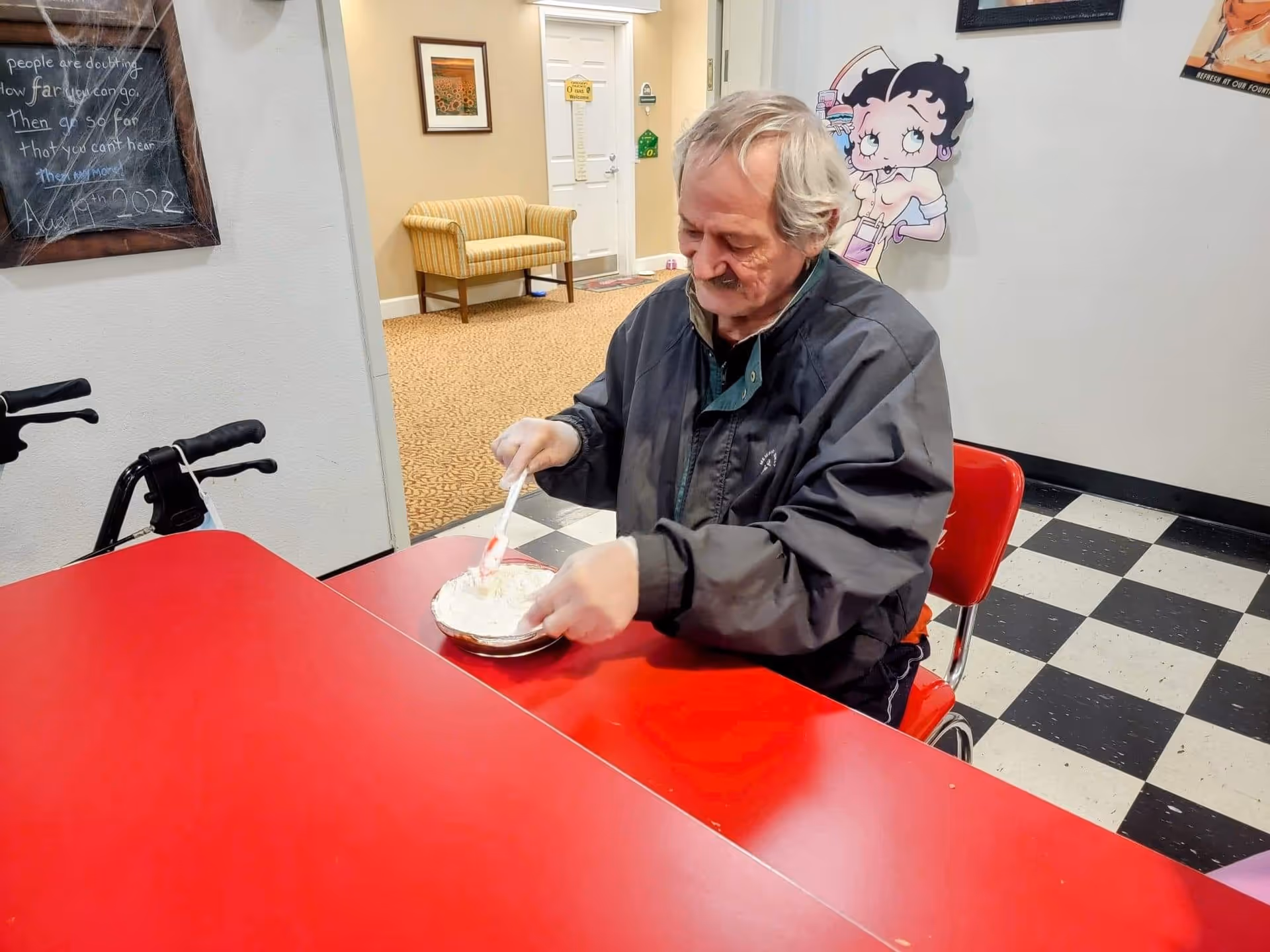 An elderly man sitting at a red table in a room with black and white checkered flooring, stirring a bowl of food with a spoon. Behind him, there is a wall with a Betty Boop poster and a hallway with a yellow striped chair and framed artwork.