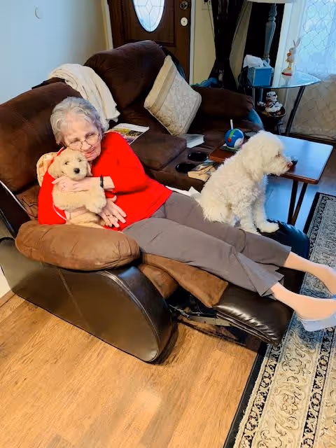 An elderly woman wearing a red sweater and gray pants is reclining on a brown leather recliner in a living room. She is hugging a small white dog, while another white dog sits on the armrest of the recliner. The room has wooden flooring, a patterned rug, a brown couch with cushions, a round side table with decorative items, and a door with a window in the background.