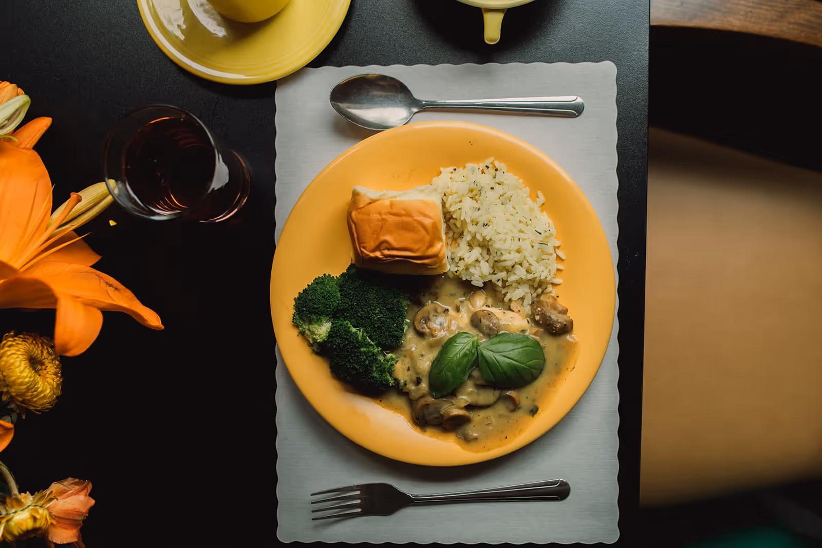 A yellow plate with a meal consisting of a bread roll, seasoned rice, broccoli, and chicken with mushroom sauce garnished with basil leaves, placed on a white placemat on a black table. A spoon and fork are set above and below the plate respectively. There is a glass of dark beverage and a yellow cup and saucer nearby, with orange flowers on the left side of the table.