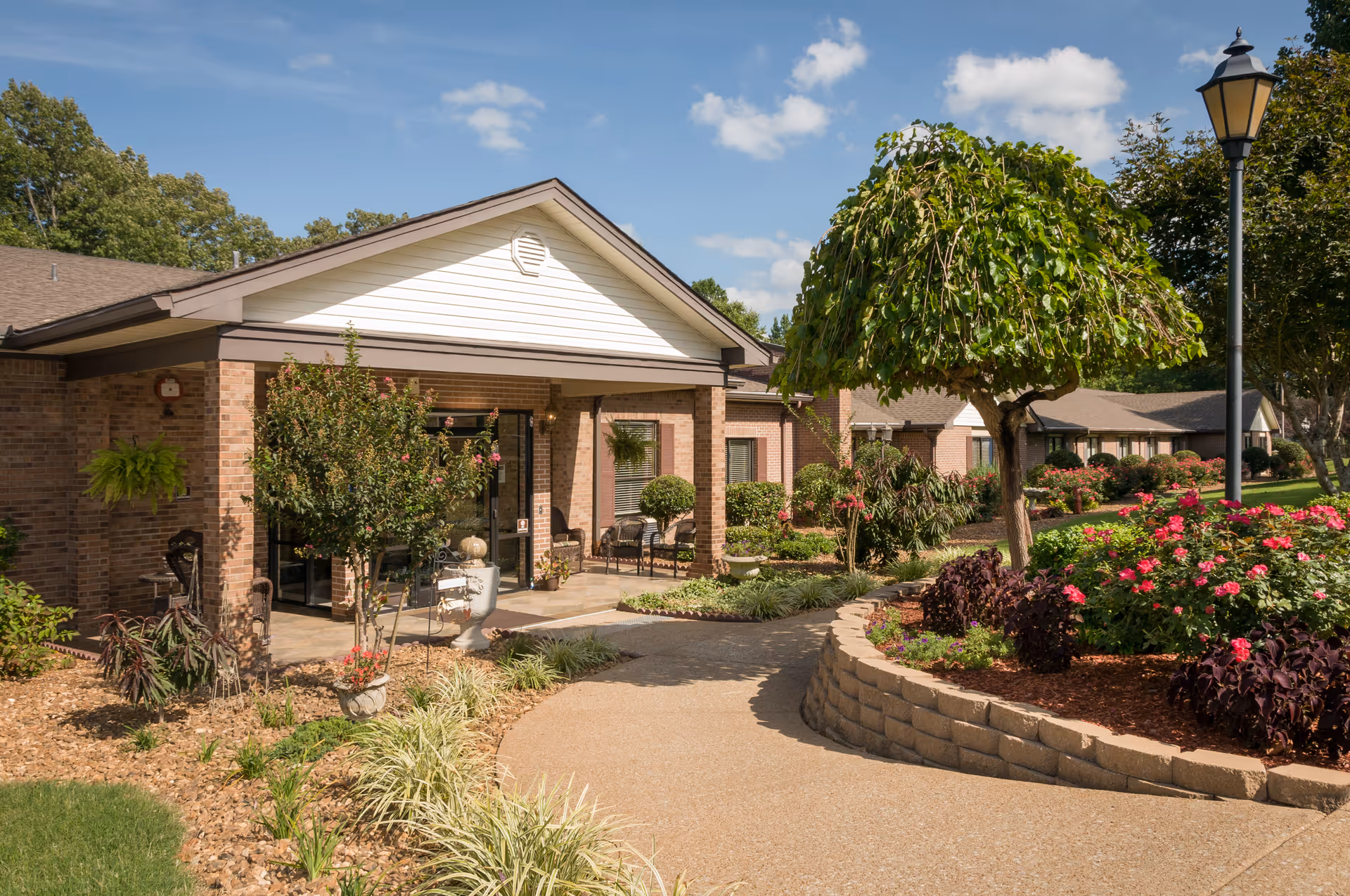 Brick single-story senior living facility entrance with a paved walkway, landscaped flower beds, and trees under a blue sky.