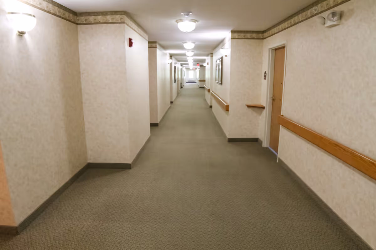 A long, well-lit hallway in a senior living facility with beige walls, carpeted floor, wooden handrails on both sides, and several doors along the corridor. Ceiling lights and wall sconces illuminate the space.