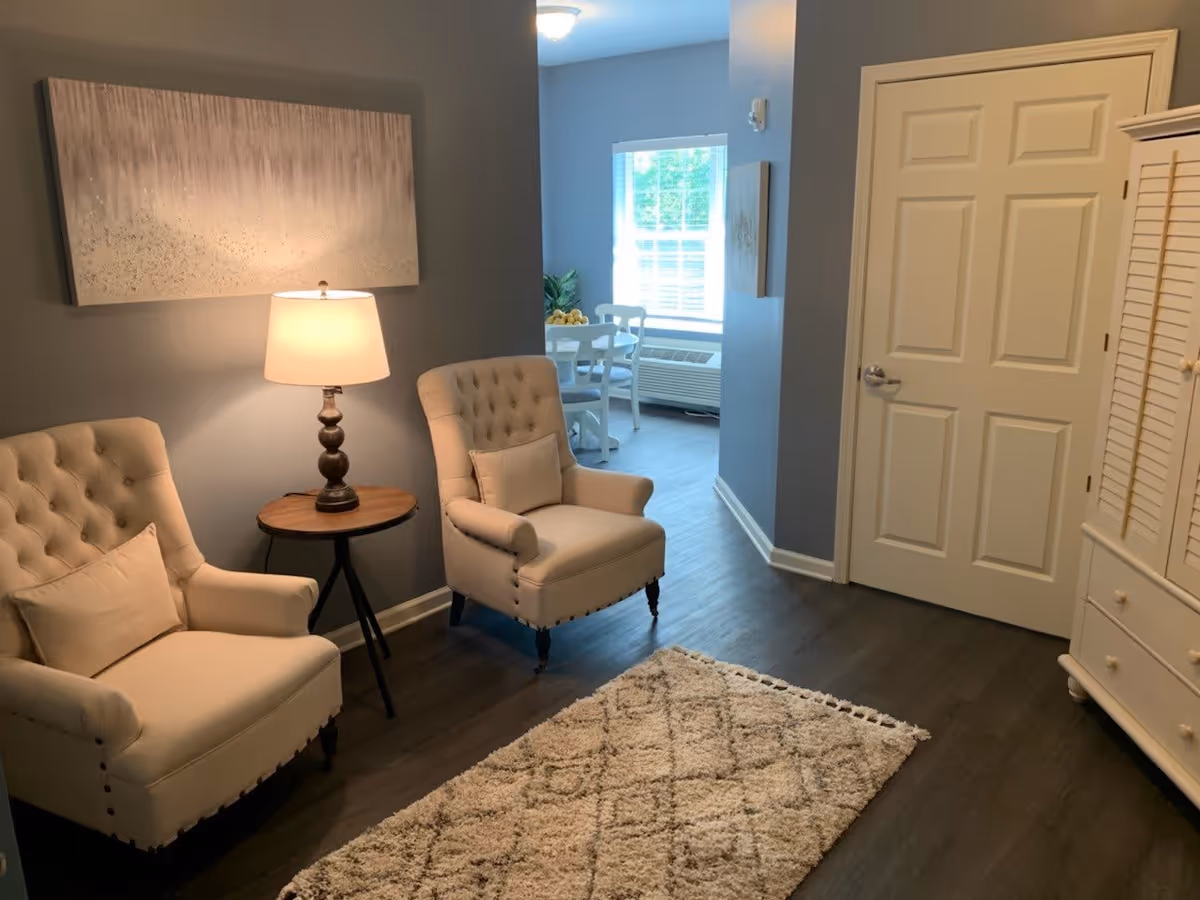 A cozy interior room with two beige upholstered armchairs with pillows, a small round wooden side table with a lamp, a textured area rug on dark wood flooring, and a white door. In the background, a dining area with a white table and chairs is visible near a window with blinds.